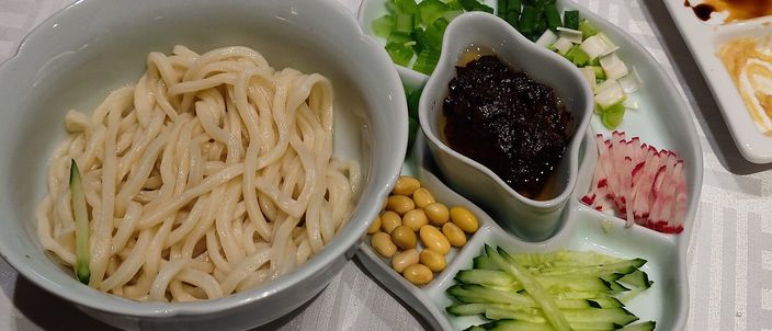 A bowl of plain thick wheat noodles served with a small dish of dark fried-soybean sauce and separate saucers of shredded cucumber, boiled soybeans, radish and greens, ready to be mixed together.