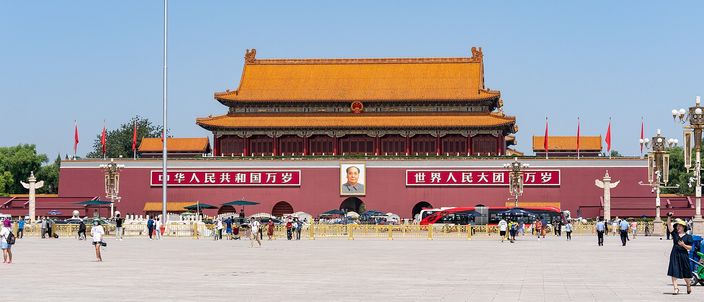 The Tiananmen gate with its red walls, portrait of Mao and two large slogan boards, yellow-tiled roof and a fluttering red flag, seen across the wide stone expanse of Tiananmen Square under a clear blue sky.