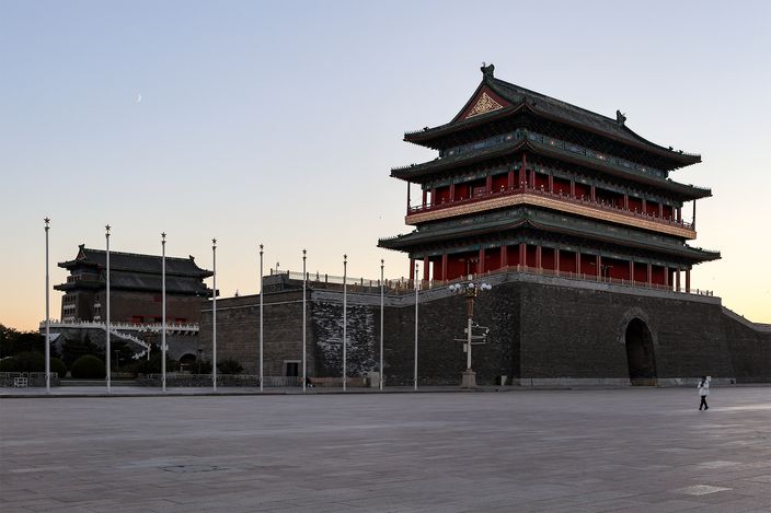 Zhengyangmen gate tower at dawn, a tall red-and-grey pavilion on a stone wall base with an arched passage, the separate lower archery tower standing across an empty paved square to the left, a few figures crossing the stone.
