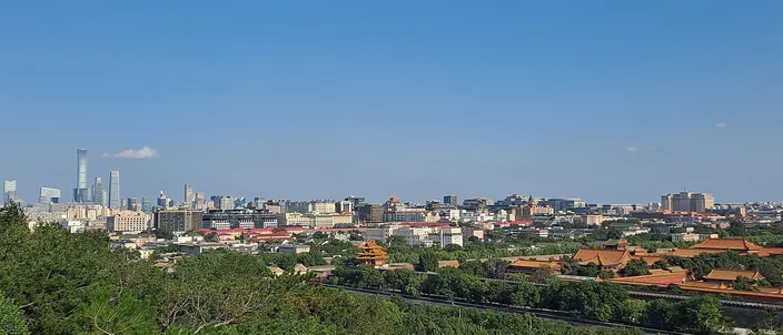 A wide daylight view over Beijing from a wooded hilltop, with the yellow-tiled roofs of the Forbidden City spread among trees on the right and the glass towers of the business district on the far horizon to the left, under a clear blue sky.