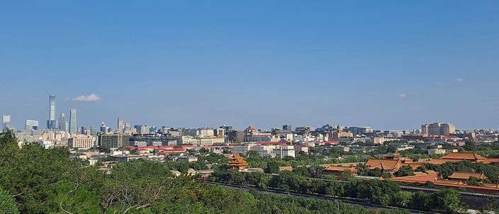 A wide daylight view over Beijing from a wooded hilltop, with the yellow-tiled roofs of the Forbidden City spread among trees on the right and the glass towers of the business district on the far horizon to the left, under a clear blue sky.