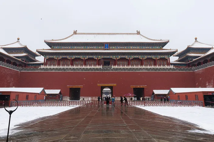 The Meridian Gate of the Forbidden City under snow: a long red wall with three arched passages, a broad dark-tiled pavilion above it and flanking wings to either side, snow lying on the roofs and on the paved forecourt, a handful of visitors crossing the wet stone.