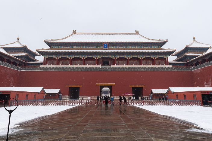 The Meridian Gate of the Forbidden City under snow: a long red wall with three arched passages, a broad dark-tiled pavilion above it and flanking wings to either side, snow lying on the roofs and on the paved forecourt, a handful of visitors crossing the wet stone.