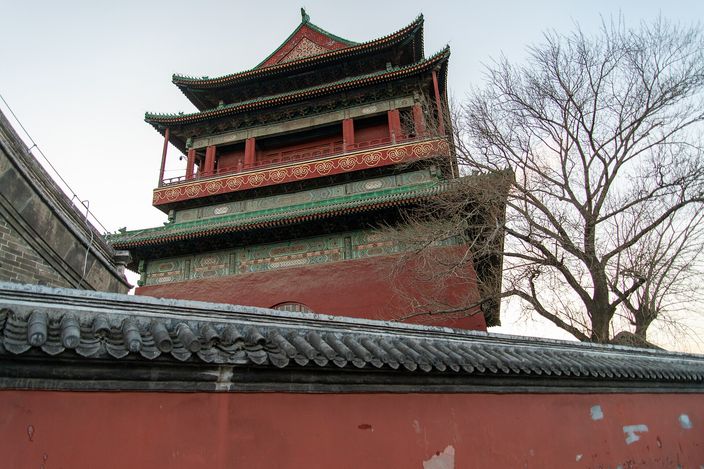 The Beijing Drum Tower seen from below and to one side in late afternoon light: a two-storey timber pavilion with green and red painted beams and upswept eaves on a massive dark red base, rising behind a grey-tiled courtyard wall with a bare tree beside it.