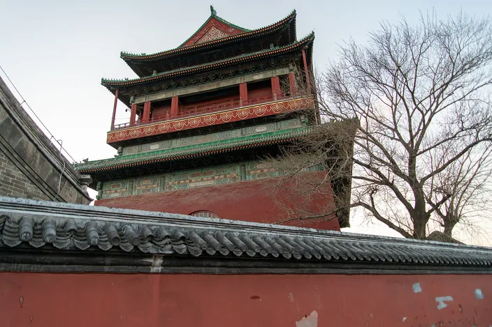 The Beijing Drum Tower seen from below and to one side in late afternoon light: a two-story timber pavilion with green and red painted beams and upswept eaves on a massive dark red base, rising behind a gray-tiled courtyard wall with a bare tree beside it.