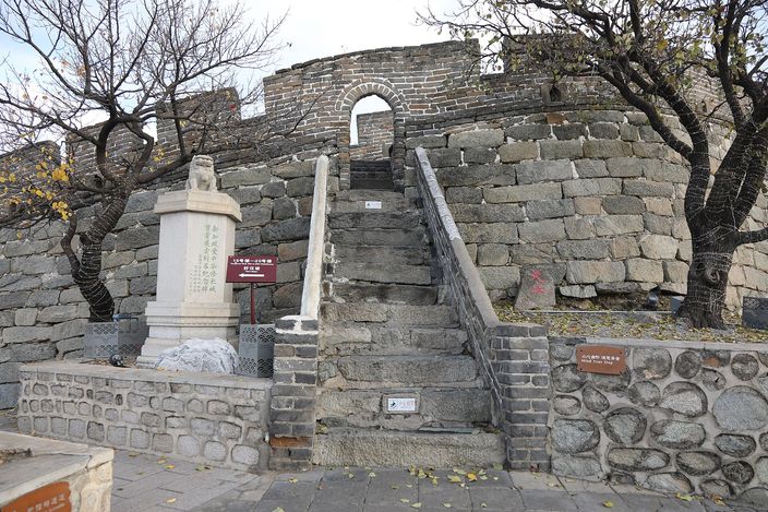 The foot of the Great Wall at Mutianyu in autumn: a stone stairway climbing between rough grey ramparts to an arched doorway in the wall base, an inscribed stone stele and a small red information board at the bottom, bare trees and fallen leaves around.
