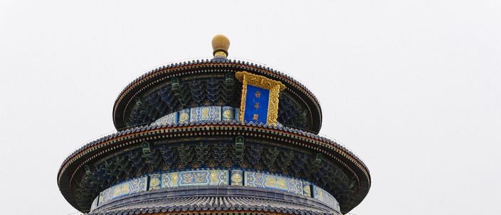 The upper tiers of the Hall of Prayer for Good Harvests at the Temple of Heaven: a gilded finial above three round roofs of dark blue tile, bands of blue, green and gold painted beamwork and a gold plaque, standing over red lattice doors against a flat white winter sky.