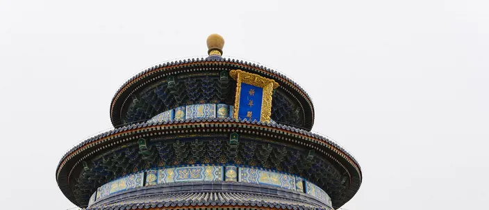 The upper tiers of the Hall of Prayer for Good Harvests at the Temple of Heaven: a gilded finial above three round roofs of dark blue tile, bands of blue, green and gold painted beamwork and a gold plaque, standing over red lattice doors against a flat white winter sky.