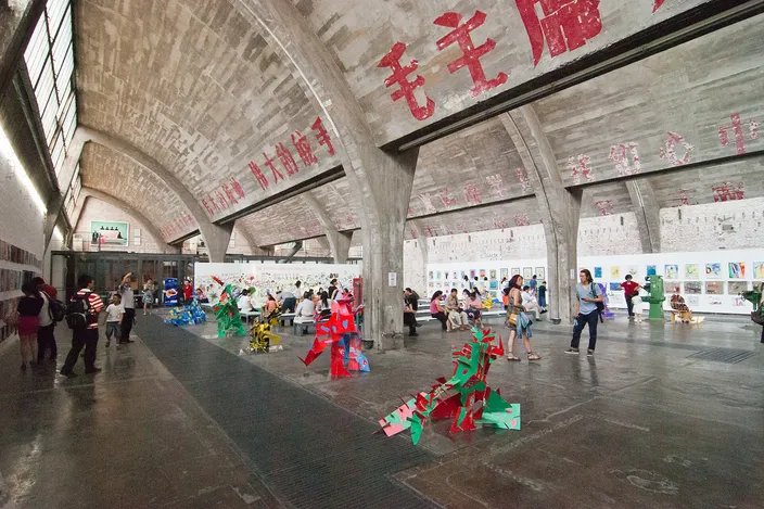 Inside a sawtooth-roofed hall of the 798 Art Zone, the original factory slogans still painted across the concrete vaults above people using the space