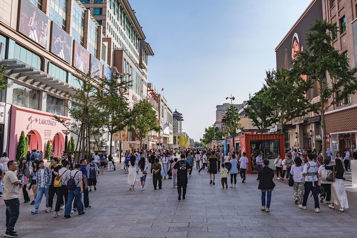 Wangfujing pedestrian shopping street in daylight, crowds strolling between department stores and trees, with a traditional decorated archway visible down the avenue