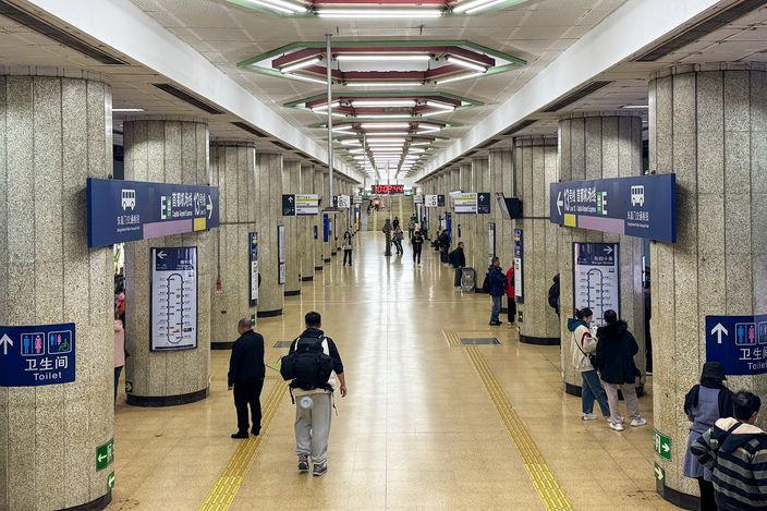 Underground platform hall of Dongzhimen Station on Beijing Subway Line 2, with wide central corridor, granite-clad pillars, bilingual blue directional signage and a few passengers walking, one with a backpack seen from behind.