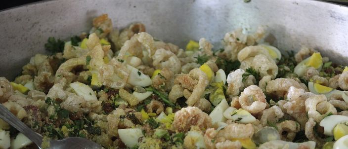 A large pan of feijão tropeiro with a serving spoon: whole brown beans tumbled with toasted cassava flour, pale pork crackling and chopped collard greens.