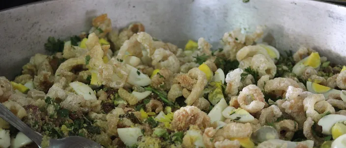 A large pan of feijão tropeiro with a serving spoon: whole brown beans tumbled with toasted cassava flour, pale pork crackling and chopped collard greens.