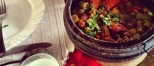 A soapstone pot of frango com quiabo — chicken braised with okra in a tomato sauce and topped with spring onion — on a Minas Gerais table beside an empty plate.