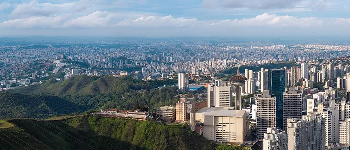 Aerial panorama of Belo Horizonte from a wooded ridge of the Serra do Curral, dense high-rise city sprawling across the valley to distant hills under a blue sky with scattered cloud