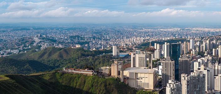 Aerial panorama of Belo Horizonte from a wooded ridge of the Serra do Curral, dense high-rise city sprawling across the valley to distant hills under a blue sky with scattered cloud
