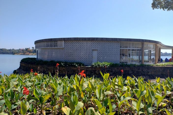 Niemeyer's Casa do Baile at Pampulha: a low circular pavilion faced in blue-and-white tiles, its flat concrete canopy curving out over a colonnaded terrace at the water's edge, with red canna lilies in the foreground and the lake and far shore behind, under a clear blue sky