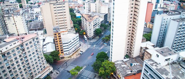 Aerial view straight down onto a downtown Belo Horizonte crossroads, where a broad avenue cuts diagonally across the street grid between pale mid-rise and high-rise blocks, with marked pedestrian crossings, street trees and blue city buses on the road below