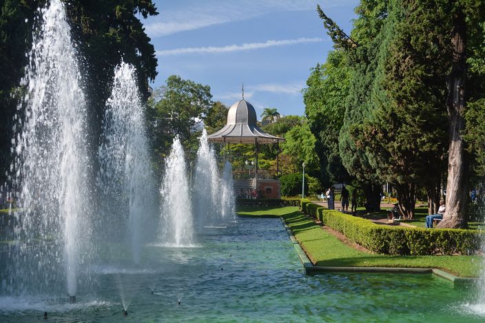 Rows of fountain jets rising from a long rectangular pool in a park, with a domed bandstand on a low terrace behind them, clipped hedges along the water and tall trees on both sides in bright sunshine.
