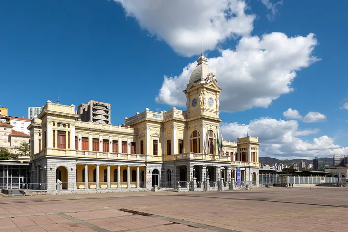 A long nineteenth-century railway terminus in cream and pale yellow, its ground floor arcaded and a central clock tower rising to a small dome, seen across the wide empty paving of its square under a blue sky with white cumulus.