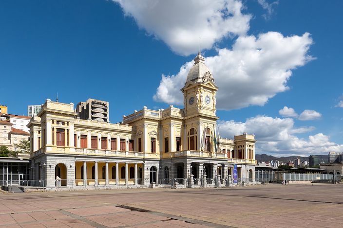 A long nineteenth-century railway terminus in cream and pale yellow, its ground floor arcaded and a central clock tower rising to a small dome, seen across the wide empty paving of its square under a blue sky with white cumulus.