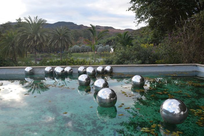 A row of mirrored steel spheres floating on a shallow green pool in a garden, with palms and shrubs beyond it and wooded hills rising behind under a pale sky.