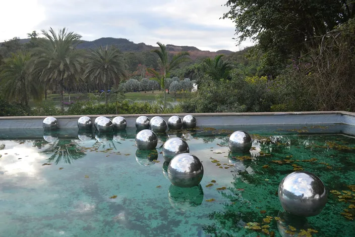 A row of mirrored steel spheres floating on a shallow green pool in a garden, with palms and shrubs beyond it and wooded hills rising behind under a pale sky.