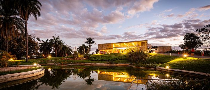 A long, low modernist pavilion of glass and pale stone glowing from within at dusk, standing on a lawn above a still pool that mirrors it, framed by palms and dark trees under a pink and grey cloudy sky.