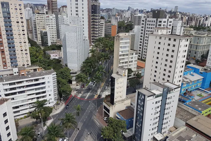 Aerial view over the Savassi/Funcionários district: dense cluster of residential high-rises with tree-lined avenues and a broad palm-lined intersection cutting through the grid, city stretching to hills on the horizon