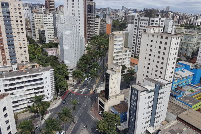 Aerial view over the Savassi/Funcionários district: dense cluster of residential high-rises with tree-lined avenues and a broad palm-lined intersection cutting through the grid, city stretching to hills on the horizon