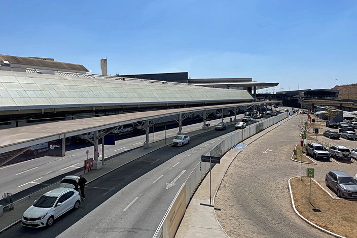 Curving canopy of Confins international airport's Terminal 2 in daylight, with the departures roadway curving past drop-off lanes, parked cars and a clear blue sky