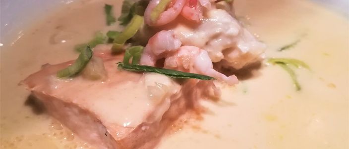 A wide white bowl of pale, creamy fish soup with a piece of poached salmon, chunks of white fish, small prawns and slivers of leek.