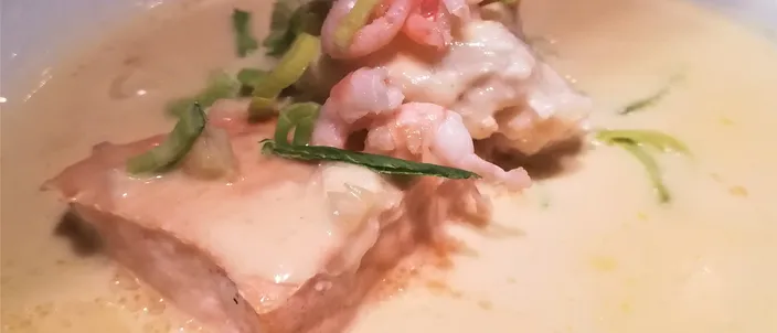 A wide white bowl of pale, creamy fish soup with a piece of poached salmon, chunks of white fish, small prawns and slivers of leek.