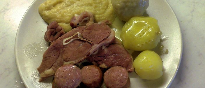 A plate holding a pale potato dumpling, a mound of yellow mashed swede, a boiled potato, slices of sausage and pieces of salted meat.