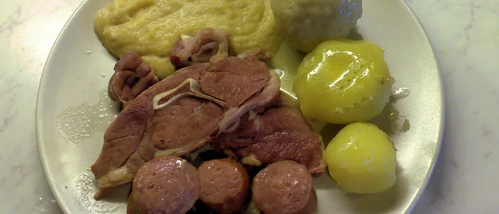 A plate holding a pale potato dumpling, a mound of yellow mashed swede, a boiled potato, slices of sausage and pieces of salted meat.