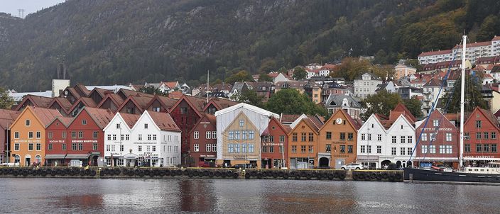 The Bryggen wharf in Bergen seen across the water: a long row of narrow wooden trading houses with pointed gables, painted ochre, red, white and cream, standing shoulder to shoulder along the quay beneath a wooded hillside of pale houses