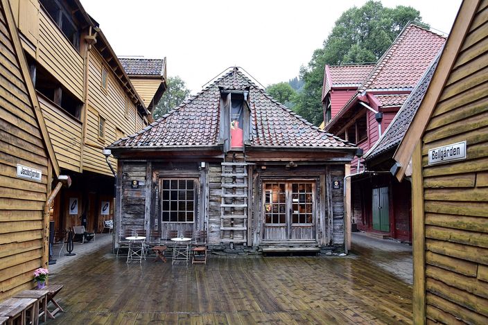 A narrow plank-floored passage between the wooden trading houses of Bryggen in Bergen, leaning weathered gables and galleries closing in on both sides, a small timber cookhouse with a tiled roof and tables outside standing at the far end, and a painted sign reading Bellgarden fixed to the boards on the right