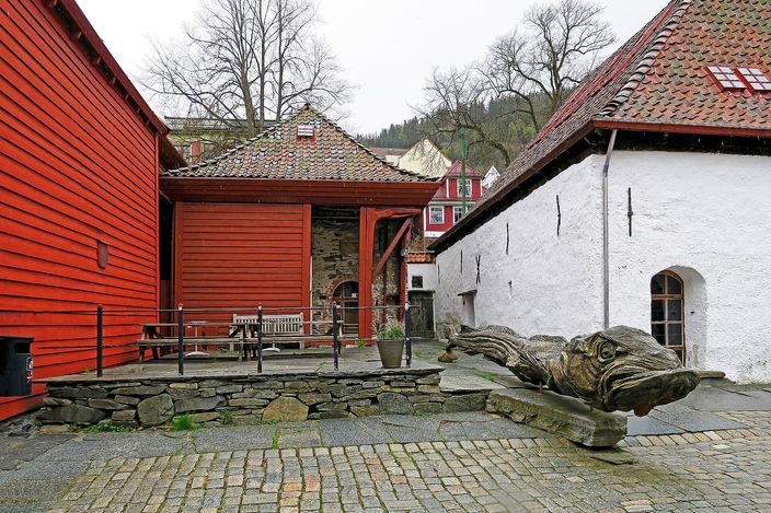A small cobbled yard at the back of the Bryggen wharf: a tall red-painted timber wall on the left, a whitewashed stone and timber building on the right, wooden benches under bare trees, and a large carved wooden fish head lying on the stones, with a wooded hillside and pale houses beyond