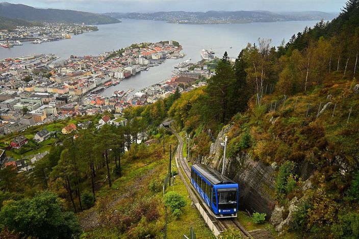 A blue funicular carriage climbing a steep single track through wooded hillside, with Bergen's harbour, its moored ships and the fjord opening out past islands to the sea far below, under a grey autumn sky