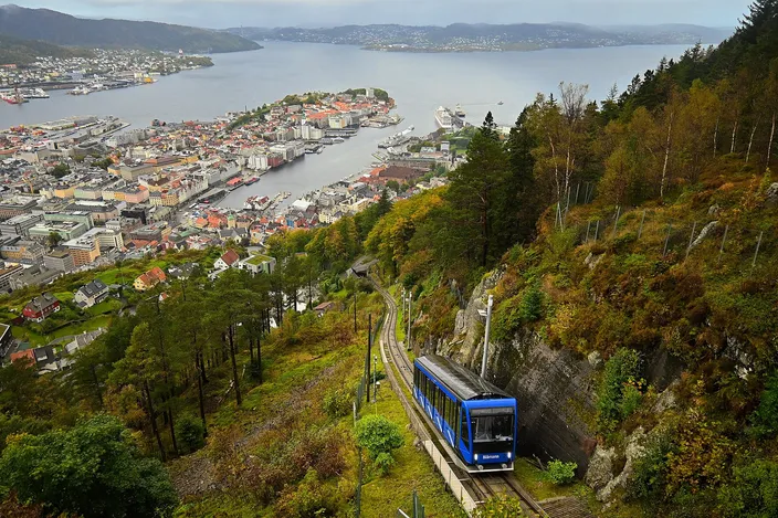A blue funicular carriage climbing a steep single track through wooded hillside, with Bergen's harbor, its moored ships and the fjord opening out past islands to the sea far below, under a gray autumn sky