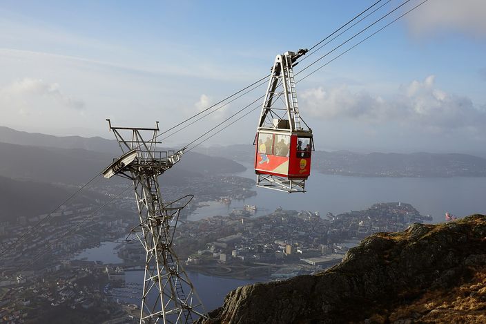 A red and white cable car gondola hanging from its cables high above Bergen, a steel lattice pylon to its left, the city with its inlets and islands spread out far below, and a bare rocky mountain slope in the bottom right corner