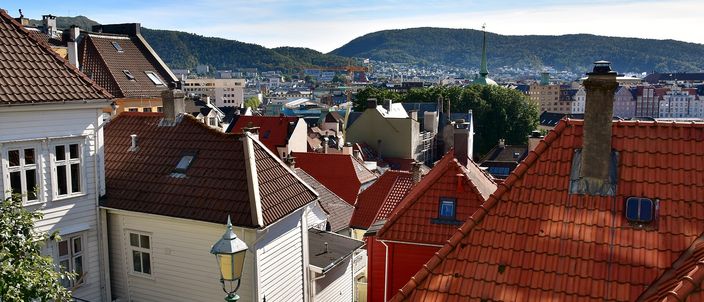 Bergen's old town seen over its rooftops: red pantiled roofs and white-painted wooden houses stepping down a slope, a black iron street lamp among them, the town and its harbour below, and a long wooded ridge of mountains behind under a blue sky