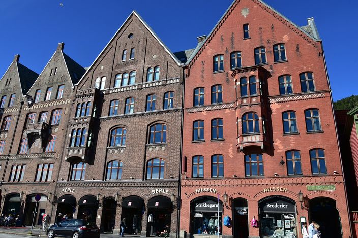 Street level on Bryggen in Bergen: tall brick warehouse buildings with stepped pointed gables and arched ground-floor shopfronts, a knitting-wool and embroidery shop among them, people walking past under a deep blue sky