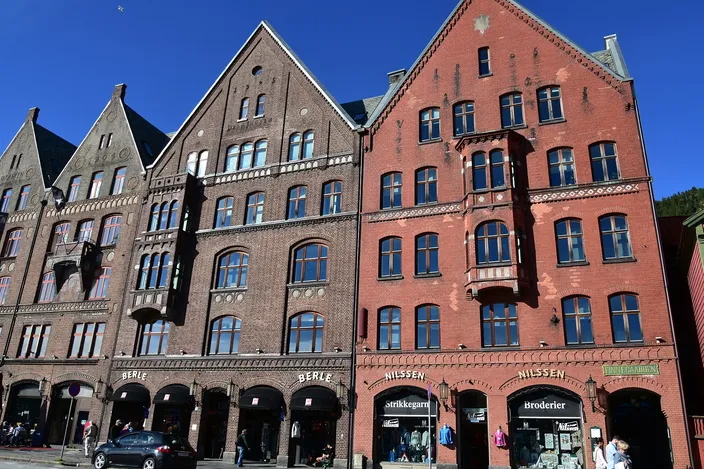 Street level on Bryggen in Bergen: tall brick warehouse buildings with stepped pointed gables and arched ground-floor shopfronts, a knitting-wool and embroidery shop among them, people walking past under a deep blue sky