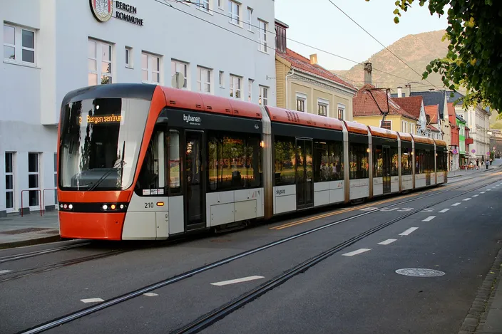 An orange and white Bybanen light rail tram on Kaigaten in central Bergen, its destination display reading Bergen sentrum, waiting on street track beside a white municipal building with a steep green mountainside rising behind the rooftops