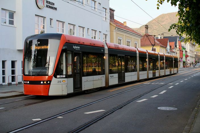 An orange and white Bybanen light rail tram on Kaigaten in central Bergen, its destination display reading Bergen sentrum, waiting on street track beside a white municipal building with a steep green mountainside rising behind the rooftops