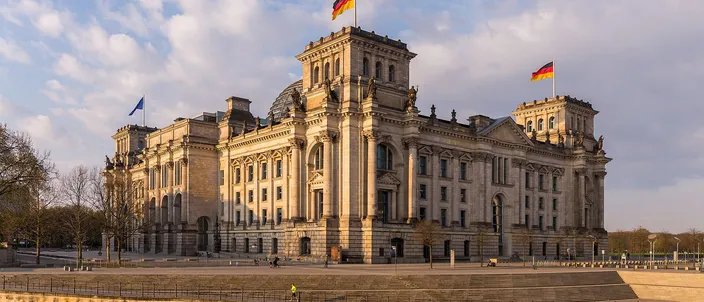 The Reichstag building in golden late-afternoon light, its neoclassical facade and glass dome mirrored in the Spree, German flags flying, bare trees under a blue-and-cloud sky.