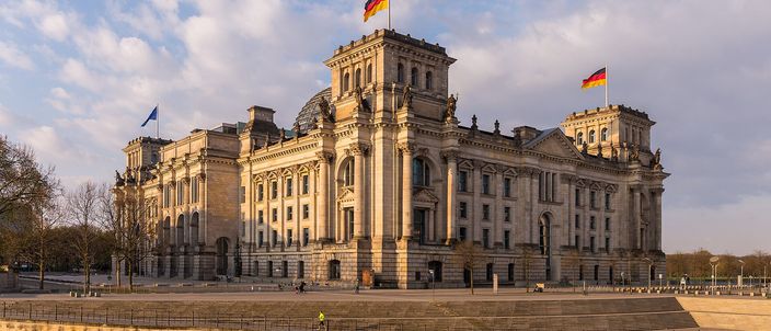 The Reichstag building in golden late-afternoon light, its neoclassical facade and glass dome mirrored in the Spree, German flags flying, bare trees under a blue-and-cloud sky.