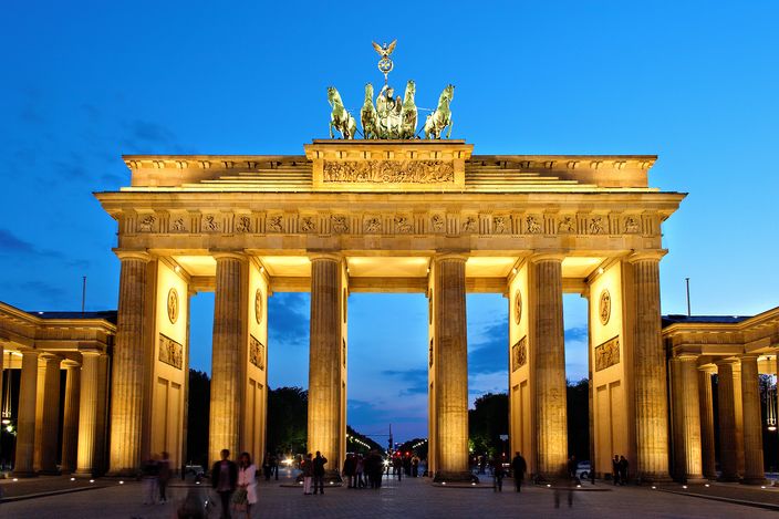 The Brandenburg Gate floodlit warm gold against a deep blue evening sky, its six columns and the bronze quadriga of four horses on top lit from below, with people crossing the paving in front of it and the tree-lined avenue visible through the central opening.