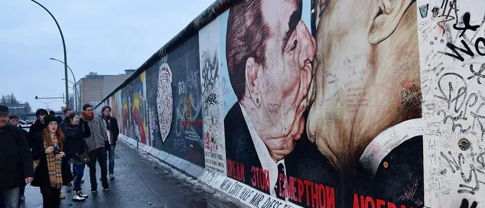 A painted stretch of the Berlin Wall at the East Side Gallery running away into the distance under a gray winter sky, the nearest segment carrying the mural of two men kissing on the mouth above red Cyrillic and hand-lettered German text, its surface covered in visitors' scribbled names; people in coats and scarves walk along the wet sidewalk beside it.